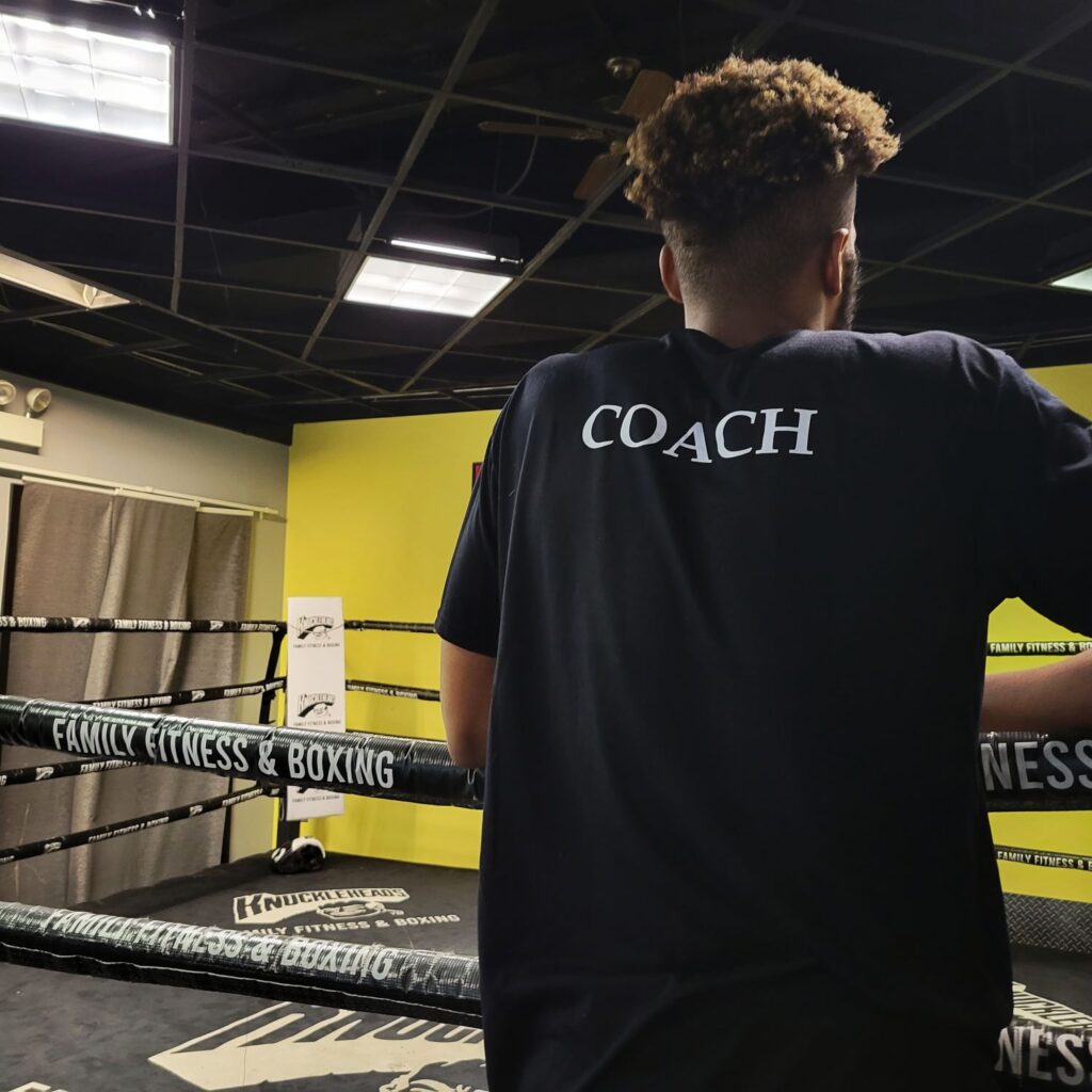 Man standing on the boxing ring with back to camera. Man is wearing a shirt that says coach.